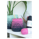 A crocheted storage basket in a pink-to-black ombre gradient sits on a white shelf next to a mint green ombre basket, with skeins of pink and black yarn and a white vase in the background.