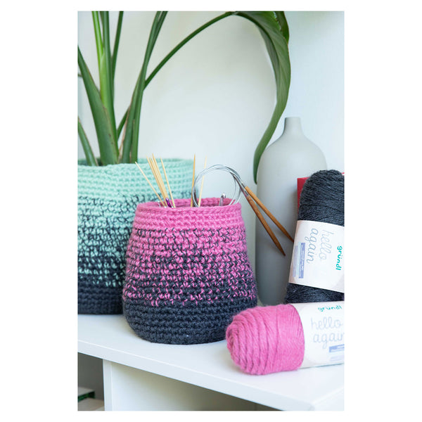 A crocheted storage basket in a pink-to-black ombre gradient sits on a white shelf next to a mint green ombre basket, with skeins of pink and black yarn and a white vase in the background.