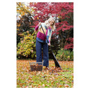 A woman in a colorful knit sweater and blue jeans rakes fallen leaves with a black rake in an outdoor yard with autumn foliage.