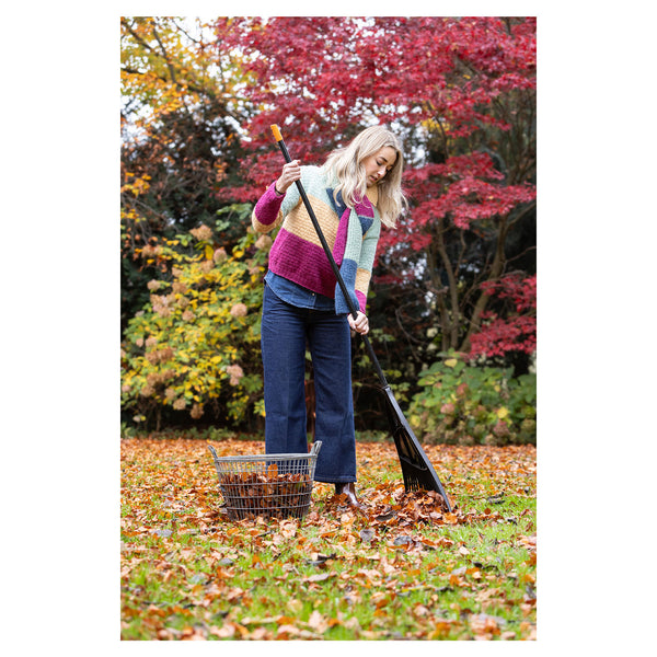A woman in a colorful knit sweater and blue jeans rakes fallen leaves with a black rake in an outdoor yard with autumn foliage.