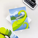 Card with a green dinosaur illustration on a blue background, surrounded by additional cards and a stamp.