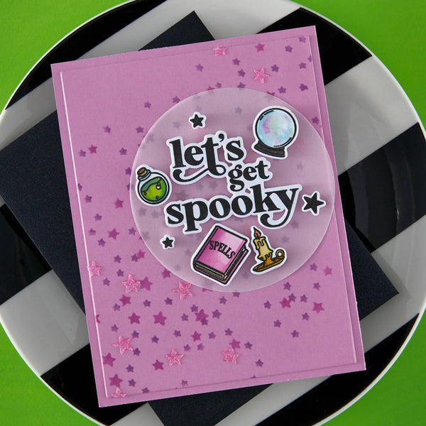A square pink card with a circular clear sticker reading 'Let's get spookty' and small spooky illustrations, placed on a black-and-white checkered plate over a bright green surface.