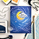 A handmade card featuring a blue starry night sky with a yellow crescent moon and a white cloud reading 'You're magical'. The card is displayed on a white surface surrounded by crafting supplies including ink pads, metal cutters, and embossing papers.