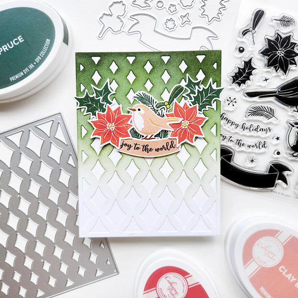 A handmade layered card featuring a robin bird, red poinsettias, and holly leaves, surrounded by crafting tools including embossing dies, clear stamps, and ink pads on a white background.