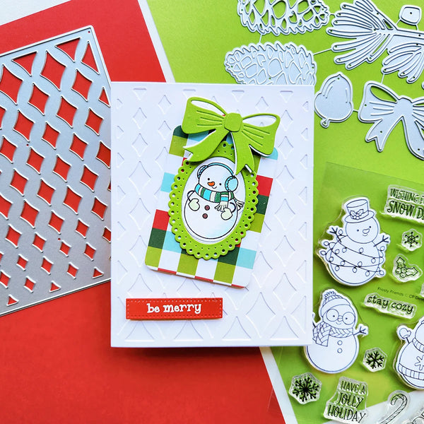 A handmade Christmas greeting card featuring a white snowman with a green bow and striped background, displayed alongside crafting dies and stamps on a red and green background.