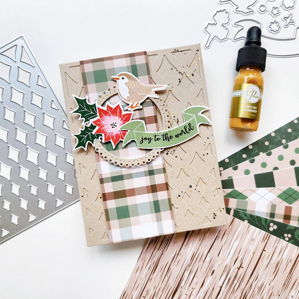 A handmade Christmas-themed card with a bird, poinsettia, holly, and 'joy to the world' banner, styled on a white surface with crafting tools and a small amber dropper bottle nearby.