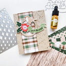 A handmade Christmas-themed card with a bird, poinsettia, holly, and 'joy to the world' banner, styled on a white surface with crafting tools and a small amber dropper bottle nearby.