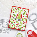 A handmade holiday greeting card with a red border, green and white holly pattern, and a central green pine tree on a white scalloped background. The card is displayed on a white surface with crafting tools and ink pads around it.