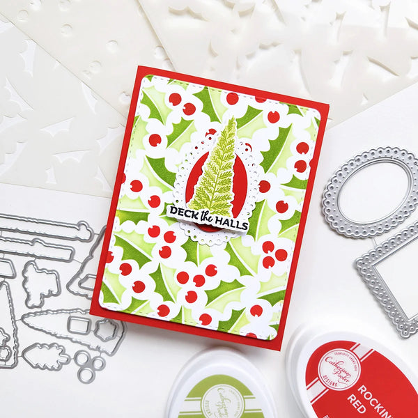 A handmade holiday greeting card with a red border, green and white holly pattern, and a central green pine tree on a white scalloped background. The card is displayed on a white surface with crafting tools and ink pads around it.