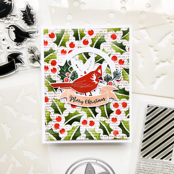 A handmade Christmas greeting card featuring a red cardinal on a patterned background of green holly leaves and red berries, with a peach banner reading 'Merry Christmas'. The card is displayed on a white background alongside crafting tools and embellishments.