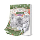 A Christmas-themed art book titled 'An Artist's Garden At Christmas' by Mark Rylance, shown standing upright with one page curled back to reveal the white interior. The cover features a festive winter village scene with snow, snowman, decorated cottage door, holly, and red berries.
