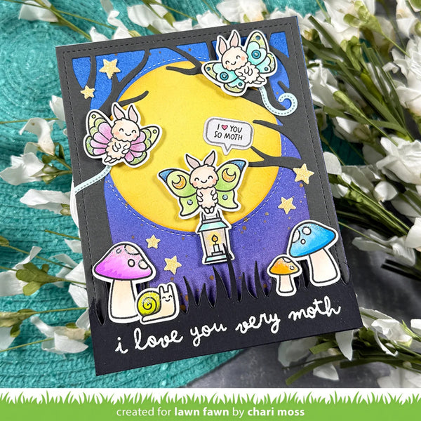A handmade greeting card featuring a whimsical moth-themed illustration, displayed on a textured teal surface with white flowers. The card has a black border and shows three colorful butterflies, a large yellow moon, a snail under a purple mushroom, and the text 'i love you very moth'.