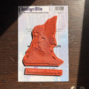 A red rubber stamp of a kneeling fairy on a light blue backing card, photographed on a dark wooden surface with sunlight casting shadows.