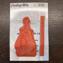 An orange-red rubber stamp and matching inked stamp block set on a printed backing card, photographed on a dark wood surface. The card shows the brand name IndigoBlu.