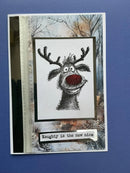 A handmade holiday greeting card featuring a black-and-white sketch of a reindeer with a red nose, set against a snowy forest background. The card has a silver glittery side panel and a small tab at the bottom that reads 'Naughty is the new nice'.