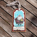 A handmade Christmas ornament shaped like a gift tag, featuring an illustration of Santa Claus holding a gift box with the text 'Be naughty, save Santa the trip.' It hangs from a white ribbon on a weathered wooden plank surface.