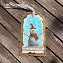 A decorative gift tag with a scalloped border hangs from a white ribbon on weathered wooden planks. The tag features an illustration of a snowman wearing a brown hat and orange scarf against a light blue background, with the text 'Baby it's cold outside.