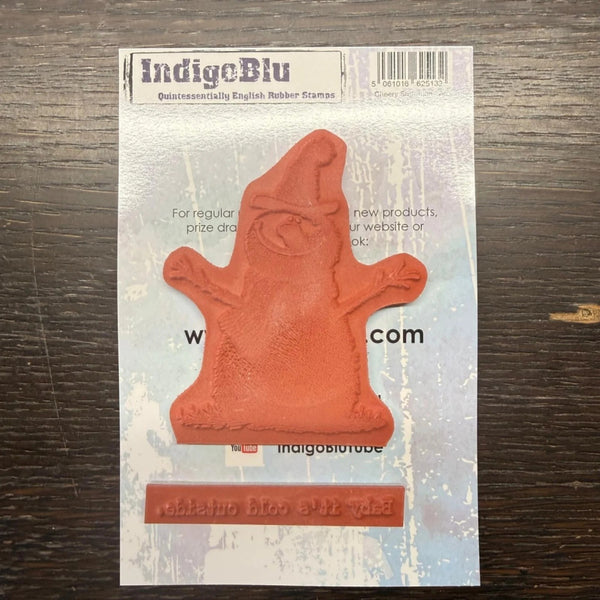 An IndigeBlu Christmas gnome rubber stamp in a terracotta orange-red color, mounted on its original printed backing card, displayed on a dark wood surface.