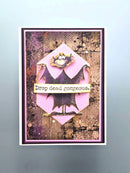 A handmade rectangular greeting card featuring a pink bat illustration with the text 'Drop dead gorgeous.' on a distressed brown and purple textured background, displayed against a light blue-gray backdrop.