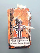 A handmade Halloween-themed paper tag or bookmark featuring a cartoon character dressed in bandages with the text 'What do you mean, it's not fancy dress.' on a mottled orange, brown, and black background, shown against a solid light blue-gray backdrop.