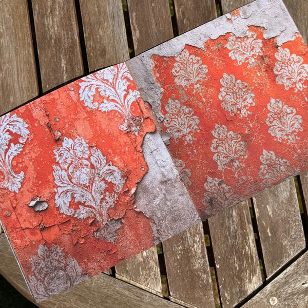 A rectangular decorative board or tray featuring a distressed red and white damask pattern, photographed diagonally on weathered wooden slats.