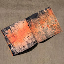 An open notebook or book with abstract painted covers in orange, black, gray, and white, photographed from above on a beige fabric surface.