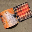 An open book or booklet laid on a textured beige fabric background, showing two printed pages with a collage-style design. The left page has orange, white, and black abstract text-like splatter graphics, while the right page features a red, orange, black, and beige harlequin diamond pattern.
