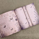 An open vintage-style journal or notebook with a dusty pink and purple gothic design, featuring illustrated butterflies, botanical sketches, and antique script, photographed on a textured beige fabric background.