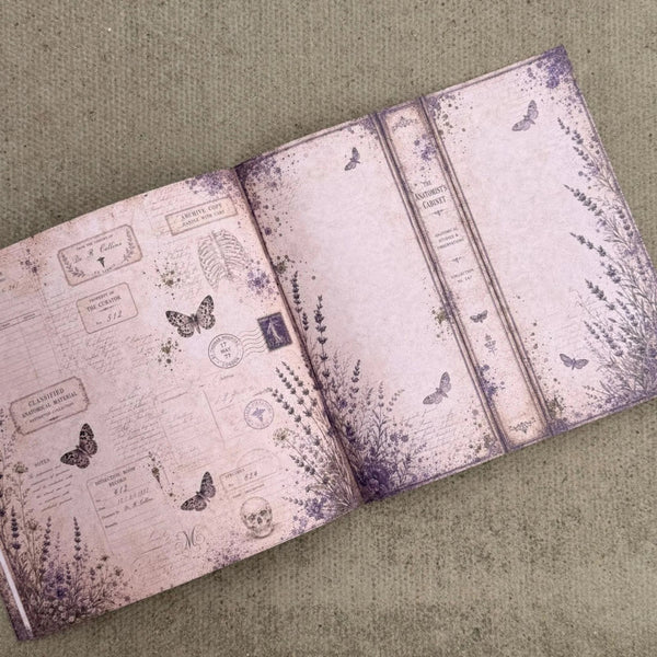 An open vintage-style journal or notebook with a dusty pink and purple gothic design, featuring illustrated butterflies, botanical sketches, and antique script, photographed on a textured beige fabric background.