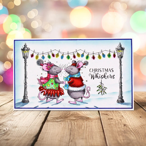 A rectangular Christmas greeting card featuring two cartoon mice in festive clothing under hanging holiday lights, with the text 'Christmas Whiskers' on the right side. The card is photographed standing upright on a wooden surface against a softly blurred multicolored bokeh background.