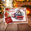 A Christmas-themed greeting card featuring a vintage steam locomotive, red poinsettias, and holiday text, displayed on a wooden surface with a warm, blurred fireplace background.