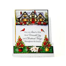 A handmade Christmas greeting card displayed on a white stand against a white background. The card features an illustration of a cozy, snow-covered cottage with red roofs and a red front door, decorated with red poinsettias and greenery.