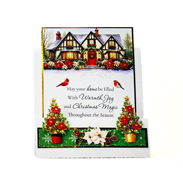 A handmade Christmas greeting card displayed on a white stand against a white background. The card features an illustration of a cozy, snow-covered cottage with red roofs and a red front door, decorated with red poinsettias and greenery.
