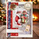 A festive handmade Christmas greeting card displayed on a wooden surface with a blurred holiday background. The card features a vintage-style illustration of Santa Claus holding a stack of wrapped presents, surrounded by red and blue patterned borders, a pinecone and holly decoration, and a hanging tag that reads 'With Love'.