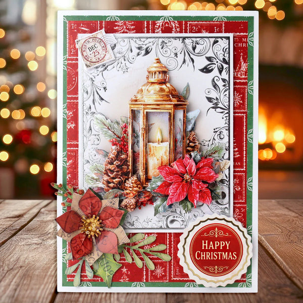 A festive handmade Christmas card displayed on a wooden surface with a blurred holiday background. The card features a red and green border, a central illustration of a gold lantern with a candle, red poinsettias, pinecones, and green foliage, and a round 'Happy Christmas' badge at the bottom right.
