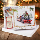 A Christmas-themed illustrated card or print displayed in a white gift box on a wooden surface, with a blurred festive background of warm lights and a Christmas tree. The artwork shows a red and black steam train with a green wreath, evergreen branches, berries, and decorative gold flourishes.