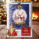 A handmade Christmas greeting card displayed on a wooden surface with a blurred festive background. The card features a snow globe design with two red deer under a full moon, decorated with holly, a gold lantern, pinecones, and a red badge that reads 'WITH LOVE AT CHRISTMAS'.