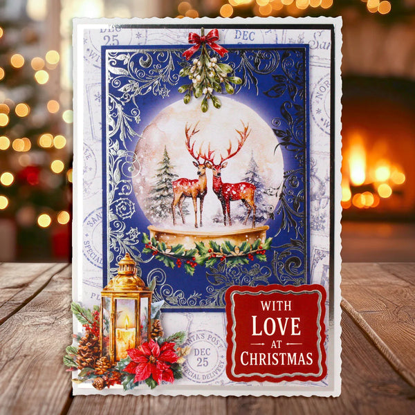 A handmade Christmas greeting card displayed on a wooden surface with a blurred festive background. The card features a snow globe design with two red deer under a full moon, decorated with holly, a gold lantern, pinecones, and a red badge that reads 'WITH LOVE AT CHRISTMAS'.