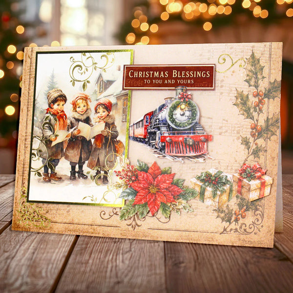 A vintage-style Christmas greeting card displayed on a wooden surface with a blurred festive background. The card features an illustration of three children singing in the snow, a red and green steam locomotive, red poinsettias, holly, wrapped gifts, and the text 'Christmas Blessings to You and Yours'.