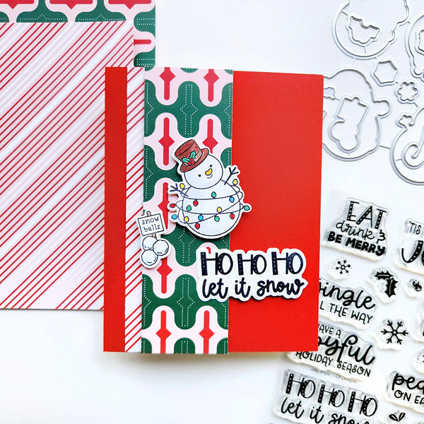 A handmade Christmas greeting card with a red and green patterned background, a red vertical panel, a die-cut snowman ornament, and the phrase 'HO HO HO let it snow' in black script. The card is styled on a white surface with patterned paper, metal cutouts, and clear stamp sheets nearby.