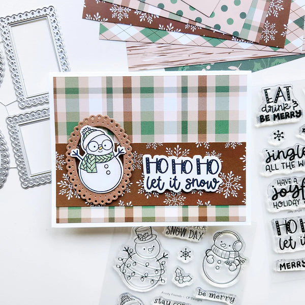 A handmade Christmas card featuring a brown and green plaid background, a scalloped oval with a snowman illustration, and the phrase 'HO HO HO let it snow' in white script. The card is displayed on a white surface with coordinating craft supplies, including patterned paper, metal frames, clear stamps, and printed phrases.
