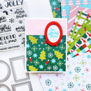 A handmade Christmas greeting card with a green patterned background, yellow Christmas trees, white snowflakes, a red scalloped oval frame, and the phrase 'Oh what fun!' in blue script. The card is styled on a white surface alongside crafting supplies, including patterned paper sheets, clear stamps, and metal die-cut frames.