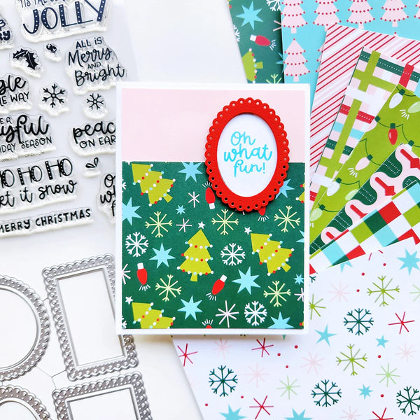 A handmade Christmas greeting card with a green patterned background, yellow Christmas trees, white snowflakes, a red scalloped oval frame, and the phrase 'Oh what fun!' in blue script. The card is styled on a white surface alongside crafting supplies, including patterned paper sheets, clear stamps, and metal die-cut frames.