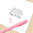 A white birthday card with printed black text and a pink handwritten signature, placed on a light wood surface with a pink pen resting across the lower part of the card.