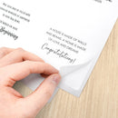 A close-up of a hand peeling back a transparent protective film from a printed card or poster, showing black text on a white background, placed on a light wood surface.
