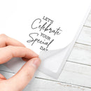 A hand peeling back a transparent protective film from a white square card printed with the phrase 'LET'S Celebrate YOUR Special DAY' in black script and block lettering, placed on a white wood surface.