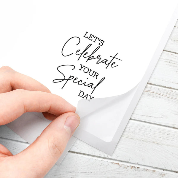 A hand peeling back a transparent protective film from a white square card printed with the phrase 'LET'S Celebrate YOUR Special DAY' in black script and block lettering, placed on a white wood surface.