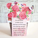 A decorative greeting card or gift box with a floral pop-up design featuring pink roses, white and purple flowers, butterflies, and a printed message on a pink background, set against a white brick wall backdrop.