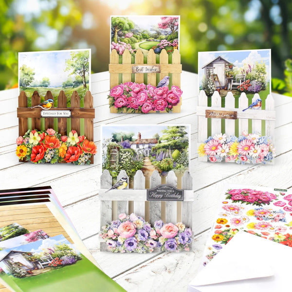 A set of decorative garden-themed cards arranged on a white wooden surface with a blurred green outdoor background. The cards feature colorful floral arrangements, picket fences, small garden scenes, and phrases like 'Welcome to My Garden,' 'Best wishes,' and 'Happy Birthday.