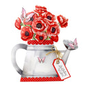 A decorative illustration of red poppy flowers in a silver watering can with pink butterflies and a red gift tag that reads Birthday Wishes, shown on a plain white background.